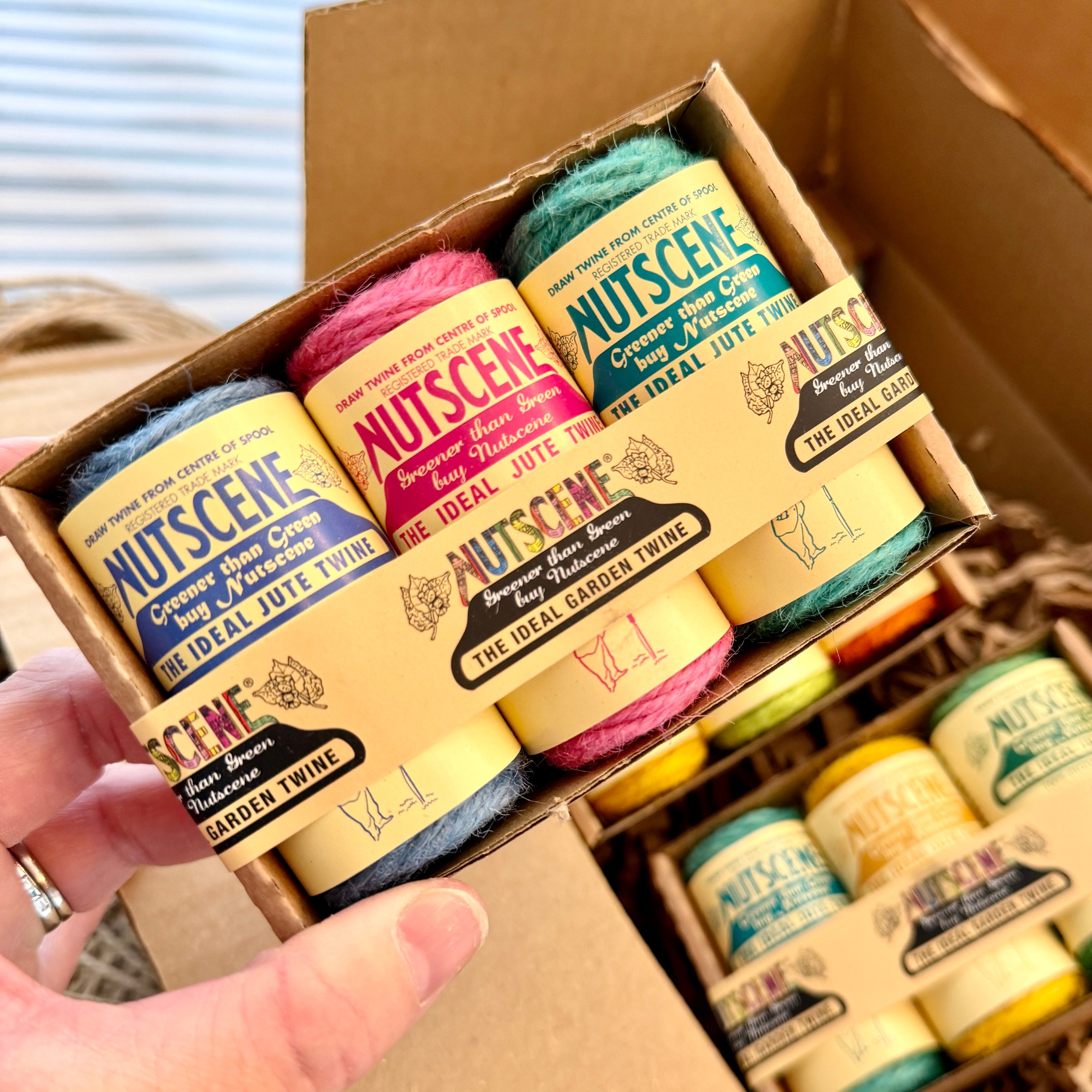 A hand holds a cardboard box containing multiple small jars of Nutscene nail polish remover. The jars are arranged in a brown paper box with shredded filler, and other boxes are visible in the background.