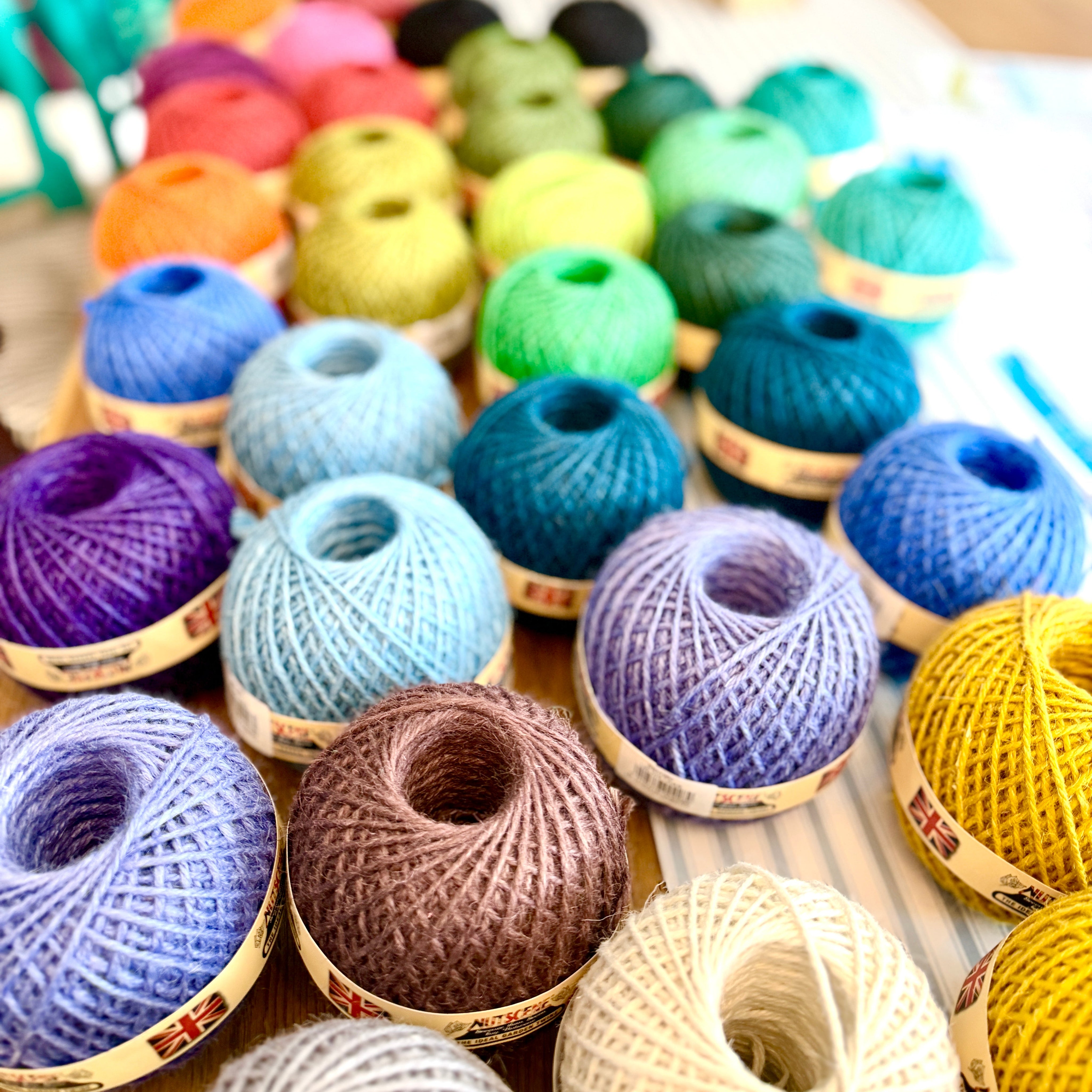 A close-up, angled view of numerous colorful skeins of yarn arranged in rows on a wooden surface. The yarns come in a wide variety of colors including purple, blue, green, yellow, brown, and cream, with some showing visible paper labels.