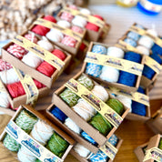 Multiple small cardboard boxes filled with colorful bakers twine bundles arranged on a wooden table. The twine comes in red, white, green, blue, and striped patterns, with paper labels wrapped around each box.