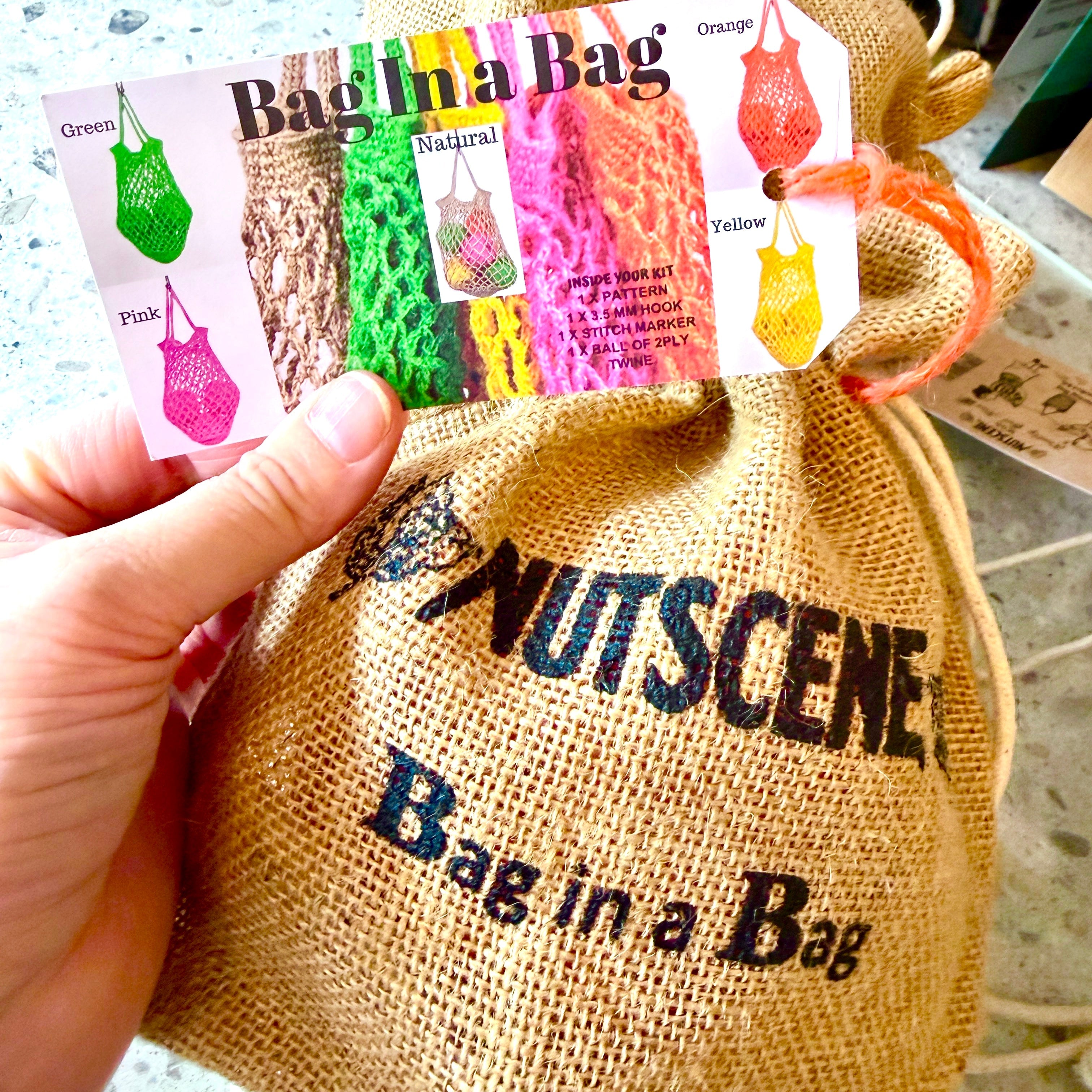 A hand holds a printed card showing color options for a 'Bag in a Bag' pouch, with a tan burlap drawstring bag printed with 'NUTSCENE Bag in a Bag' in the foreground.