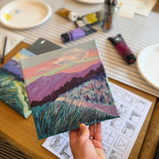 A hand holds a small square canvas painting of a colorful landscape with purple mountains and a pink sky, set on a wooden table surrounded by art supplies like paint tubes, brushes, and paper plates.