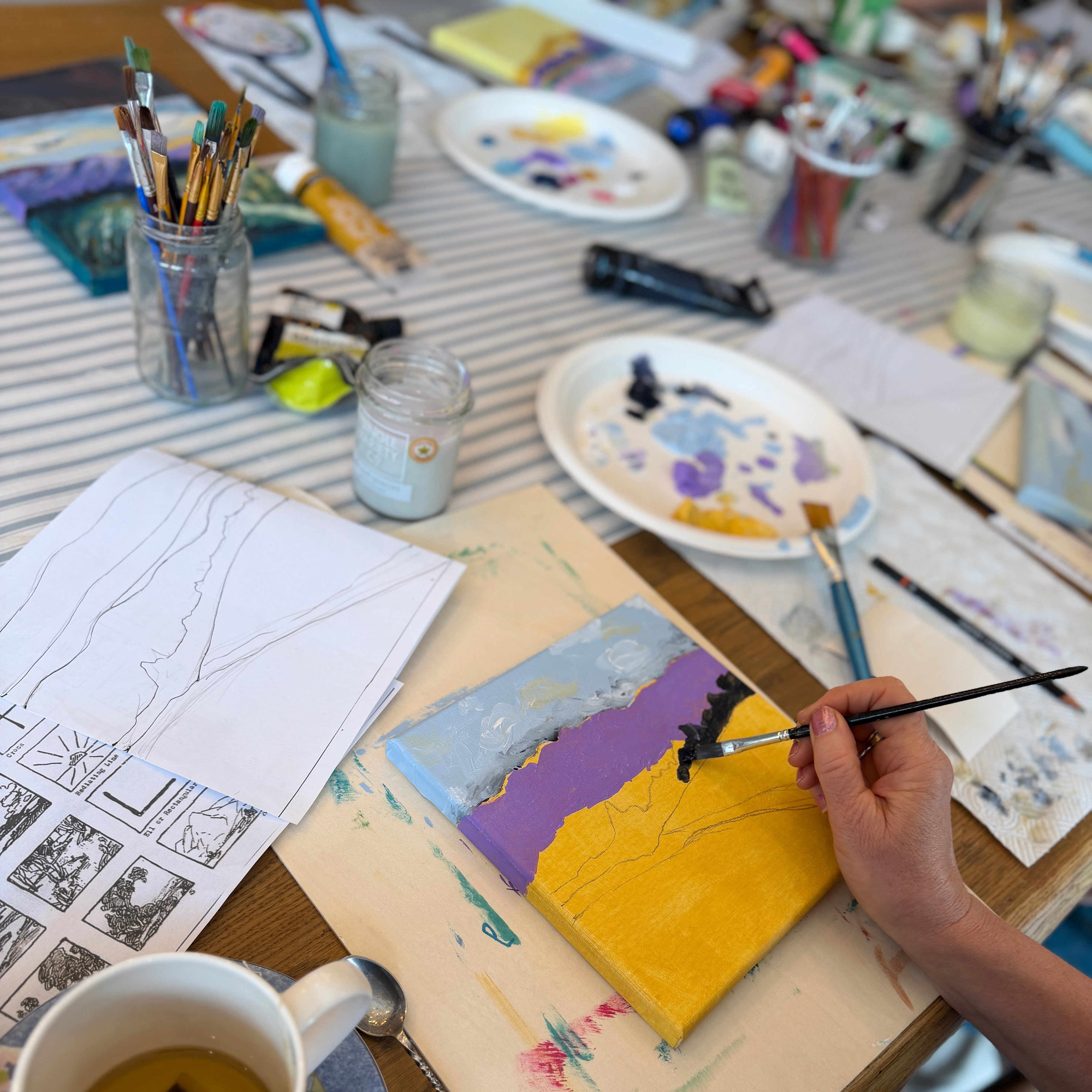 A person's hand holding a paintbrush, painting a small canvas with yellow, purple, and blue sections. The scene is set on a wooden table covered with art supplies, including paint palettes, brushes, sketch papers, and a teacup on a saucer.