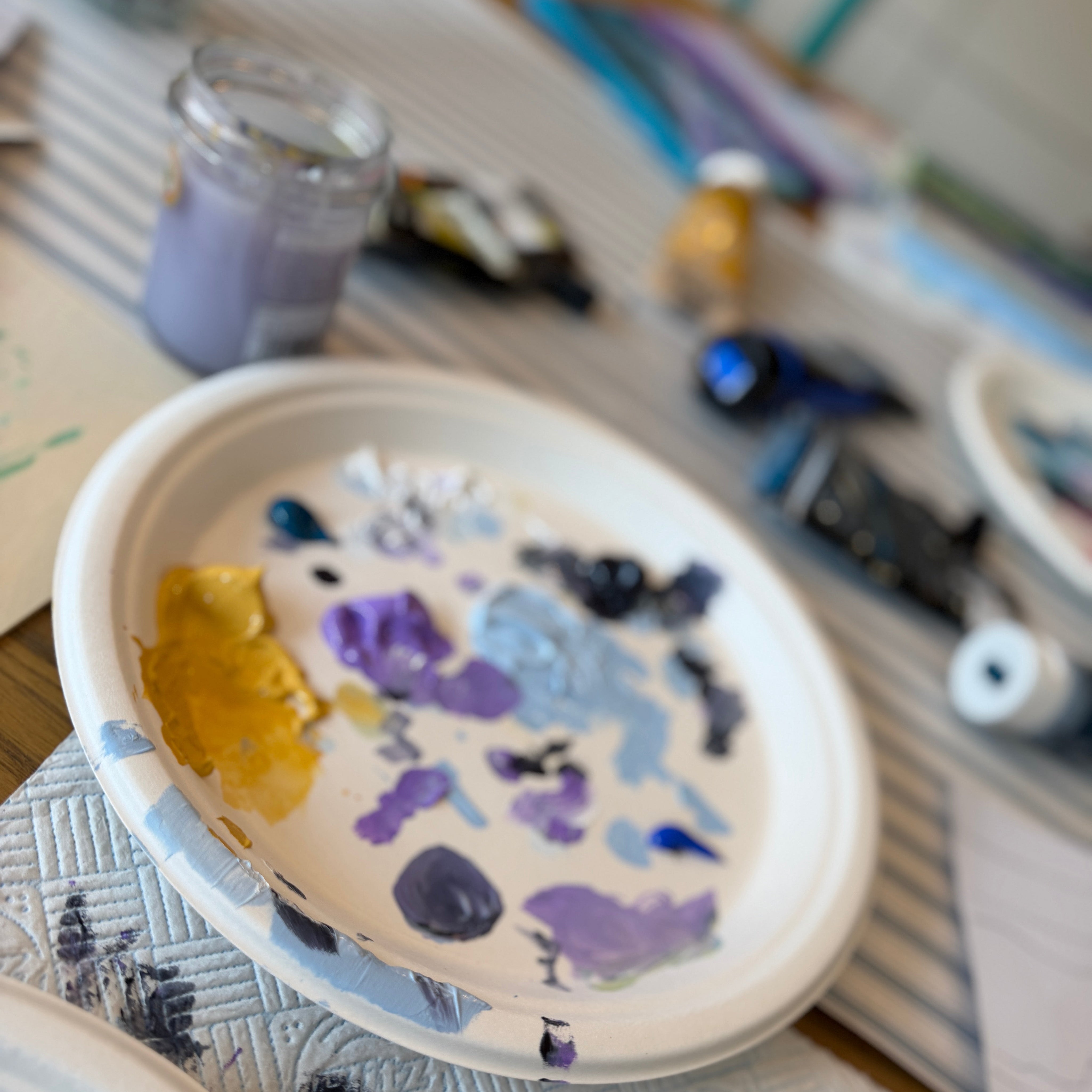 A white paper plate acting as a paint palette with various colors of paint (yellow, purple, blue, black, white) spread inside, set on a patterned white napkin on a wooden table surrounded by art supplies.