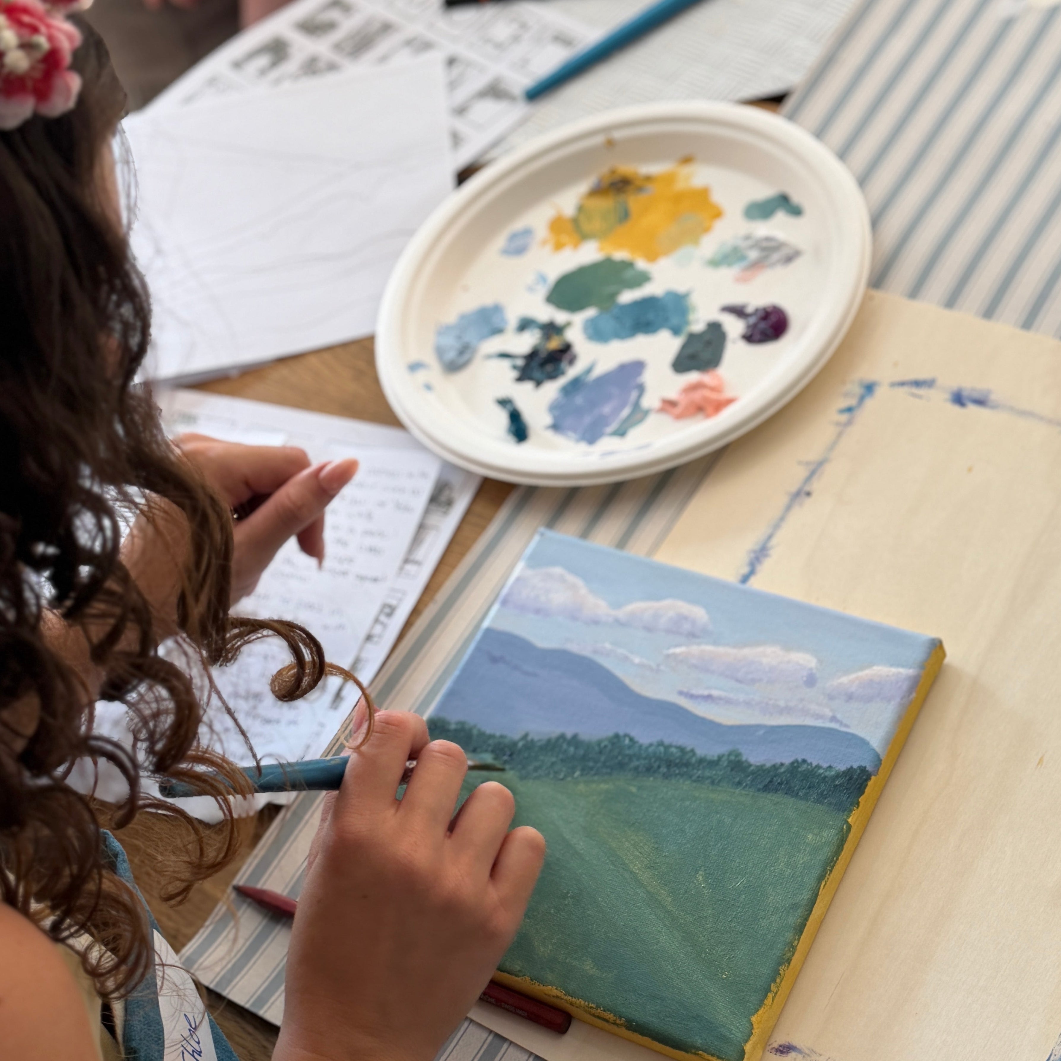 A child painting a landscape scene on a small canvas at a table with an artist palette, paintbrushes, and other art supplies.