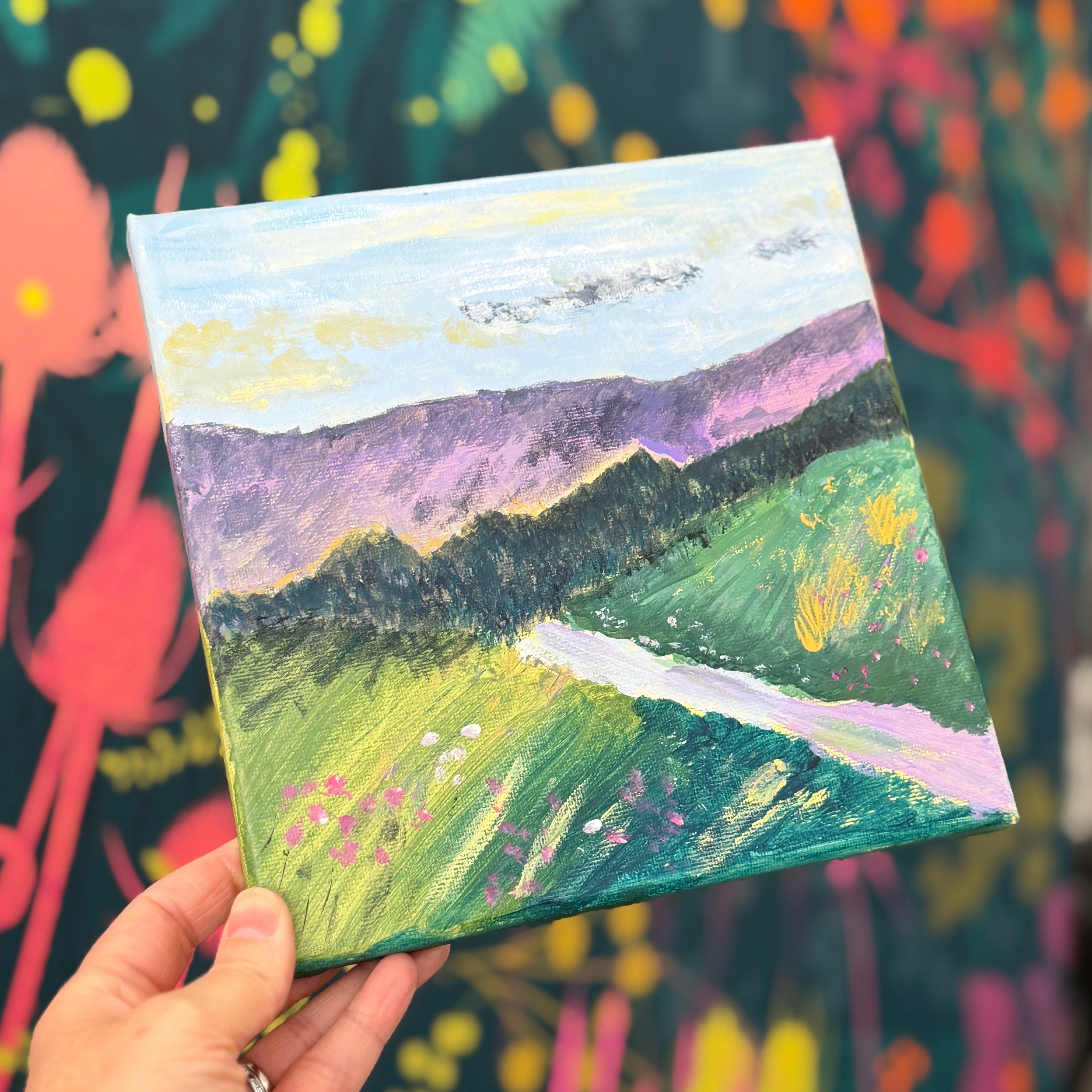 A hand holds a small square canvas painting depicting a colorful landscape with green hills, purple mountains, and a winding white road. The background features a large, vibrant abstract floral or botanical painting in a multicolor palette.