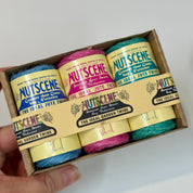 A hand holds a small cardboard box containing three spools of Nutscene cotton jute twine in blue, pink, and teal, with the labels clearly visible.