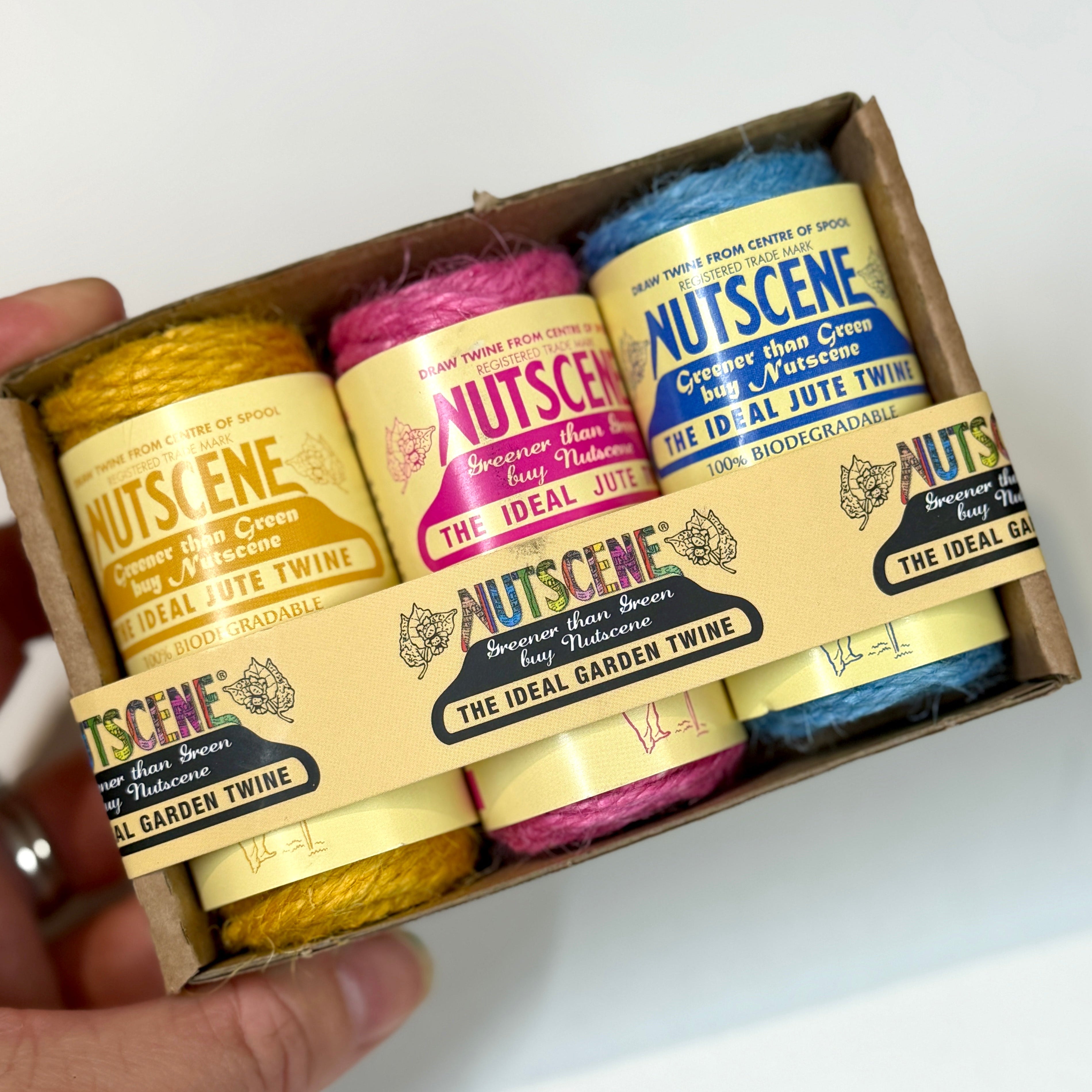 A hand holding a small cardboard box containing three skeins of jute twine in yellow, pink, and blue. The labels read Nutscene and The Ideal Garden Twine.