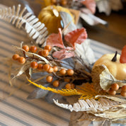 An autumn-themed decorative centerpiece with small pumpkins, dried leaves, orange berries, and textured botanical elements arranged on a striped surface.