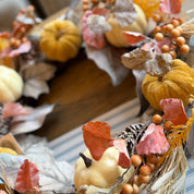 An autumn-themed decorative wreath featuring faux pumpkins, dried leaves, pinecones, and orange berries, displayed on a wooden surface.