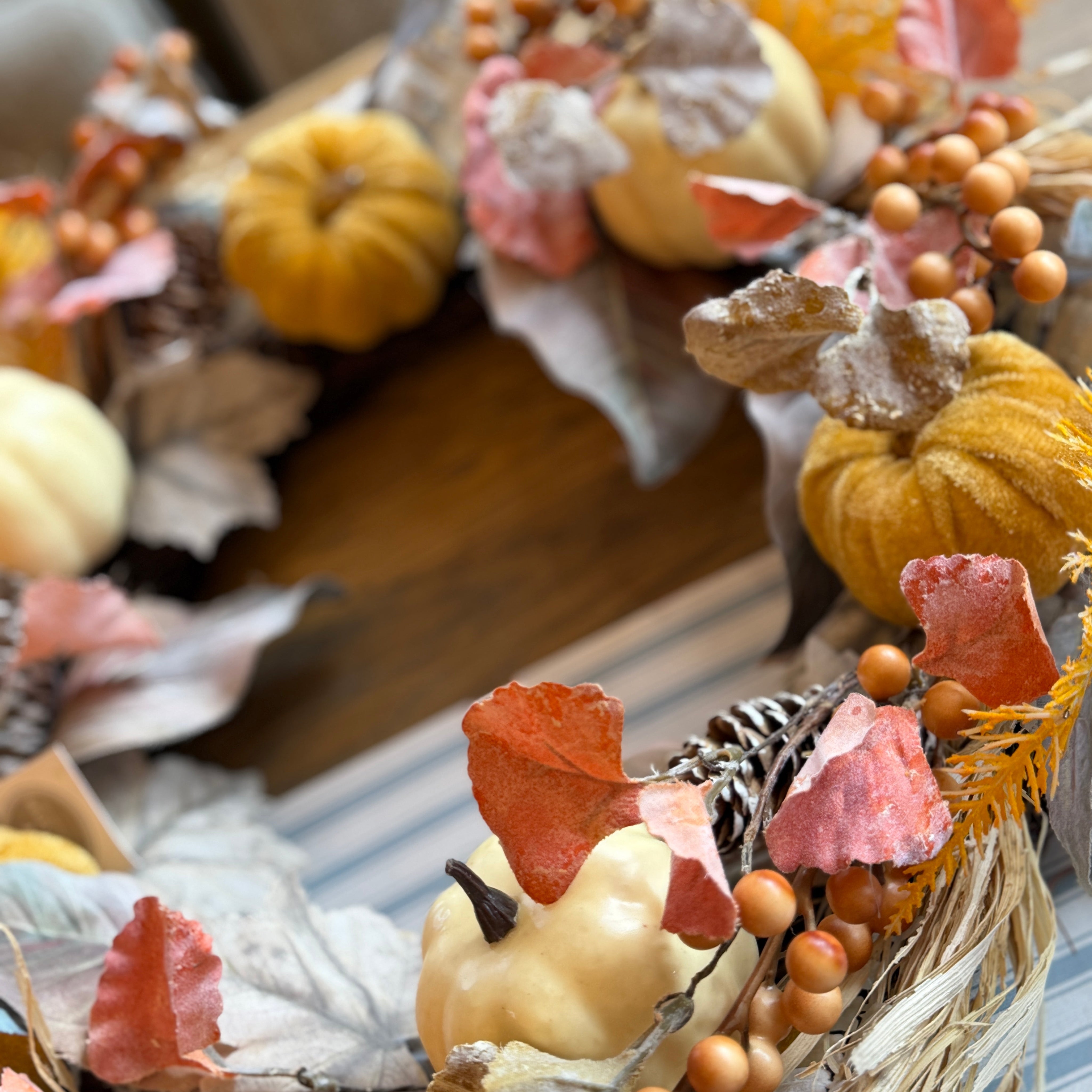 An autumn-themed decorative wreath featuring faux pumpkins, dried leaves, pinecones, and orange berries, displayed on a wooden surface.