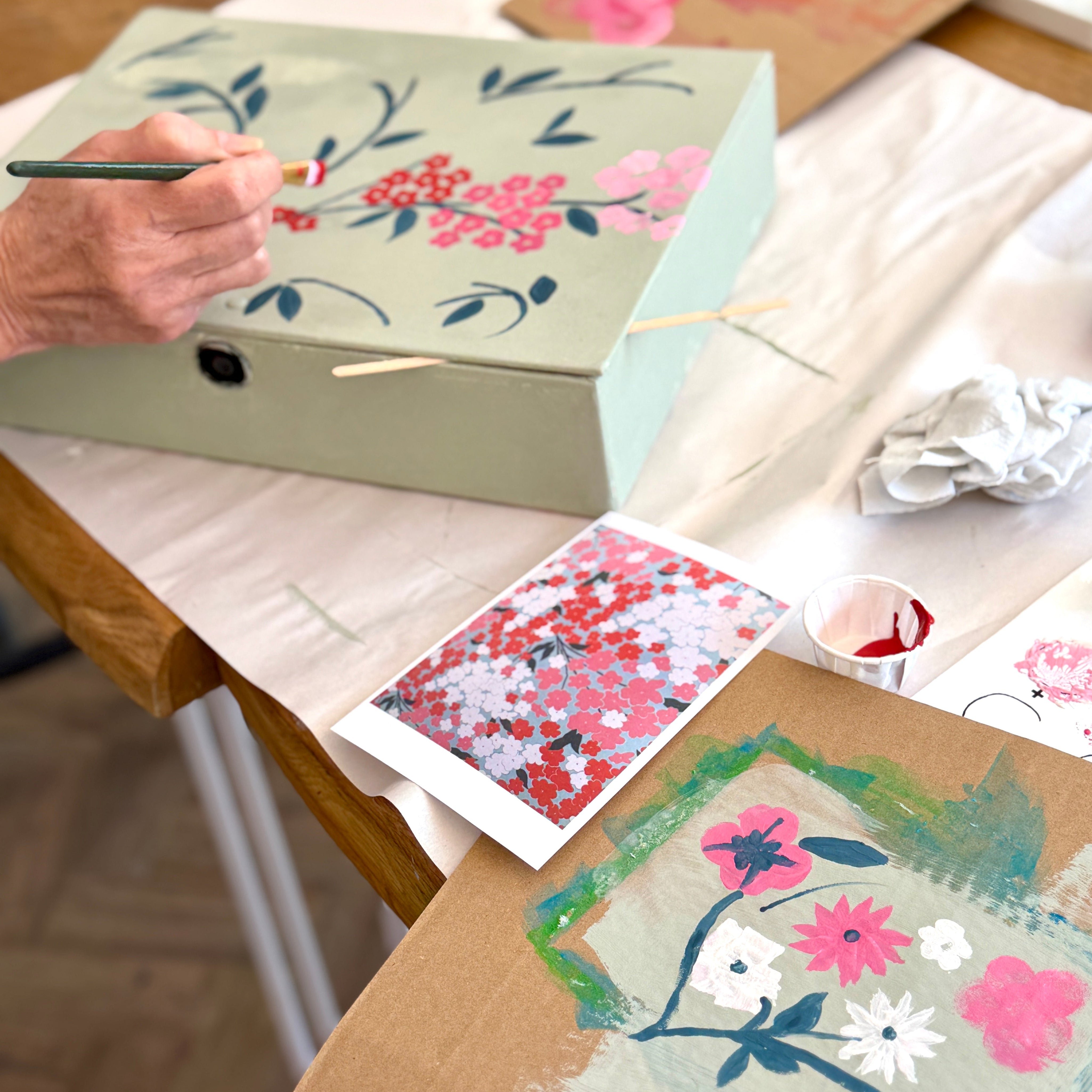 A hand holds a paintbrush, painting floral designs onto a light sage green box. The scene is set on a wooden table covered with a white sheet, surrounded by art supplies, painted samples, and other decorated boxes in a warm, creative workshop environment.