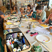 A group of people seated around a long table in a craft studio or art shop, engaged in painting and drawing activities. The table is covered with art supplies, including paint tubes, brushes, palettes, paper, and markers.