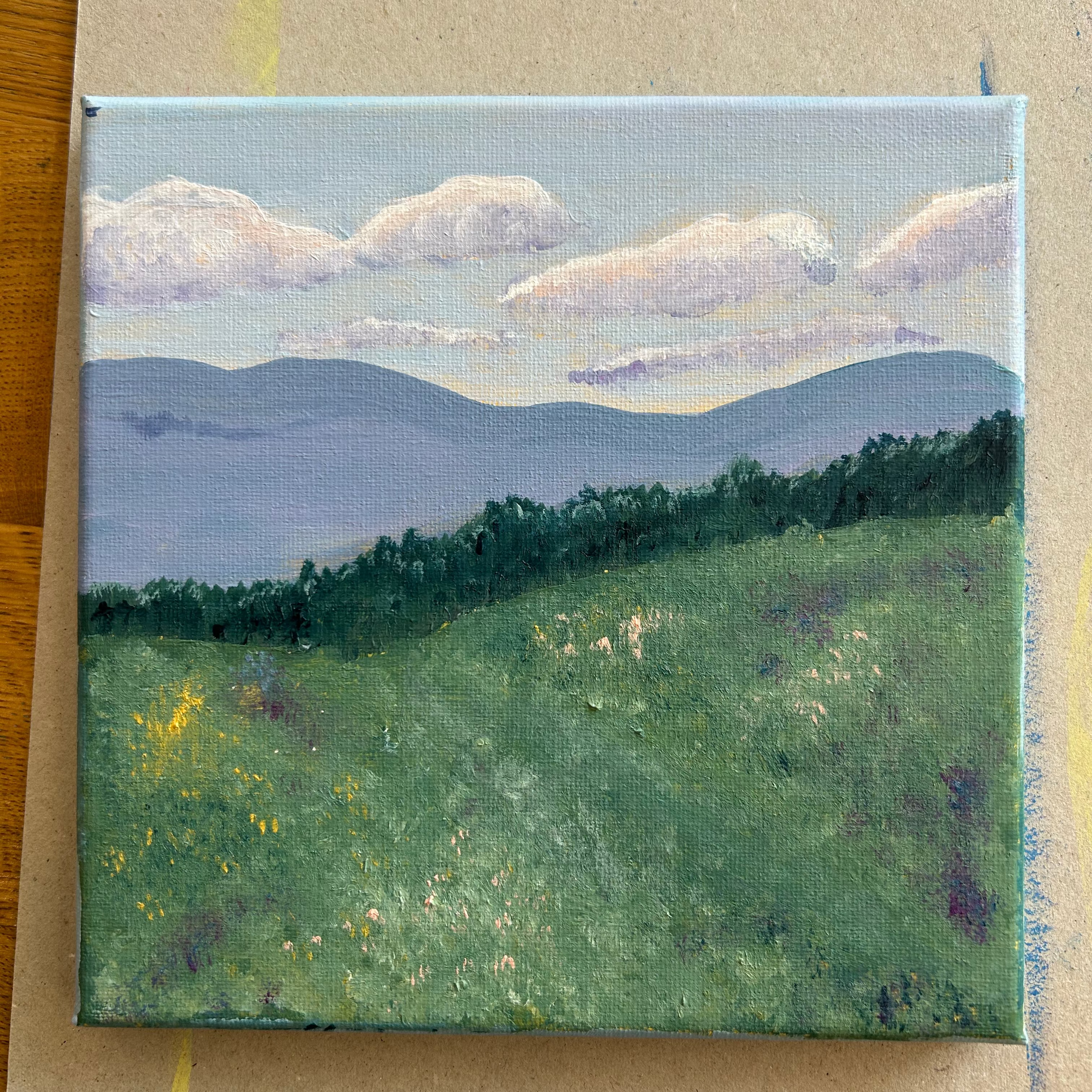 A small square canvas painting of a landscape with green fields, dark tree lines, layered blue mountains, and a pale sky with white clouds, photographed on a tan cardboard surface.