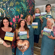 A group of seven people, including a baby, wearing blue aprons stand in front of a colorful floral wall, each holding a small landscape painting. A heart-shaped balloon reading 'True Bride' is held in the center.