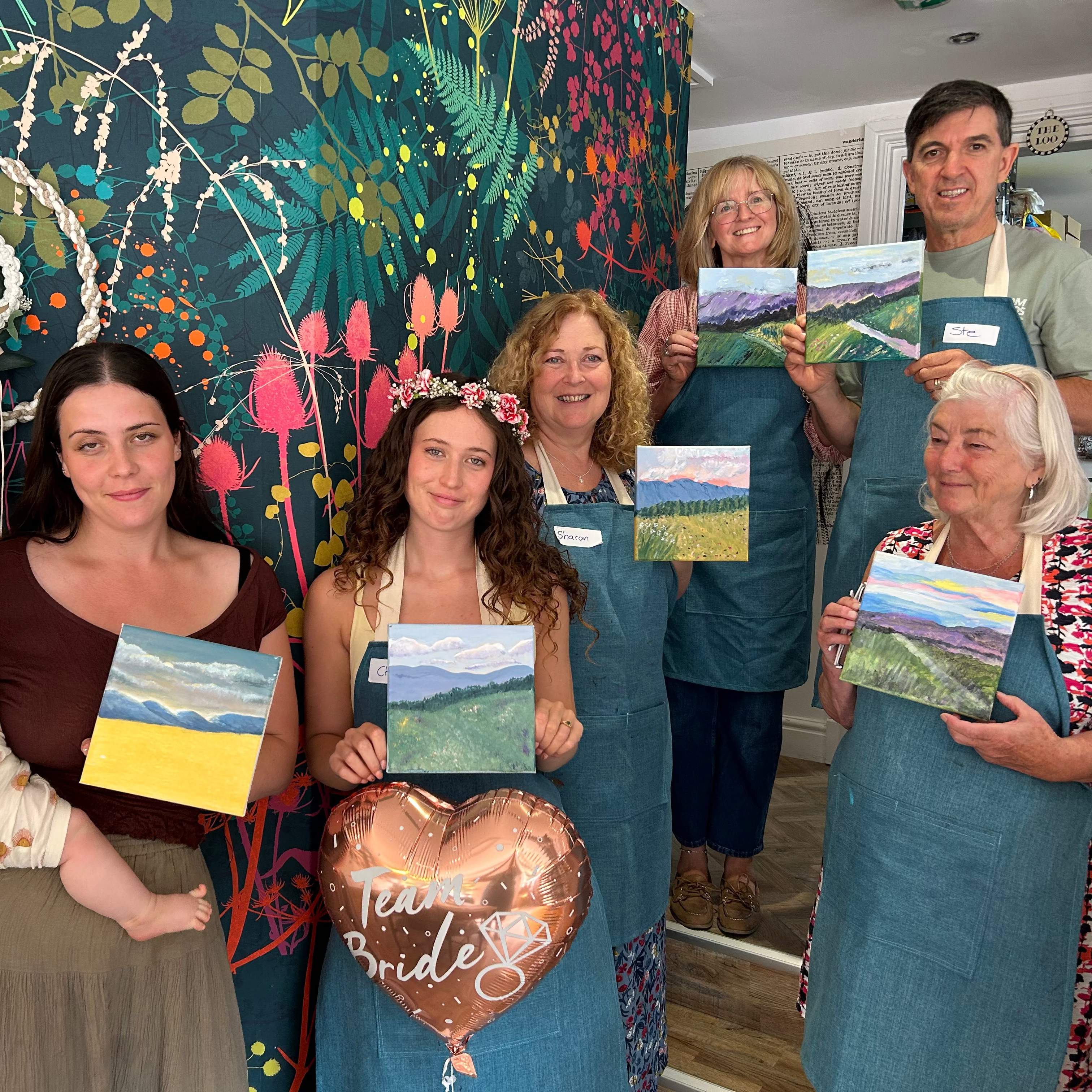 A group of seven people, including a baby, wearing blue aprons stand in front of a colorful floral wall, each holding a small landscape painting. A heart-shaped balloon reading 'True Bride' is held in the center.