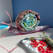 A handmade quilted snail doll with a pink and purple body, a blue and green patterned shell, and pink yarn antennae, displayed on a white surface against a blue background.