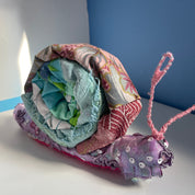 A handmade fabric snail decoration made from patterned and solid colored cloth layers, with pink yarn antennae and sequins on the body, resting on a white surface against a blue and white background.