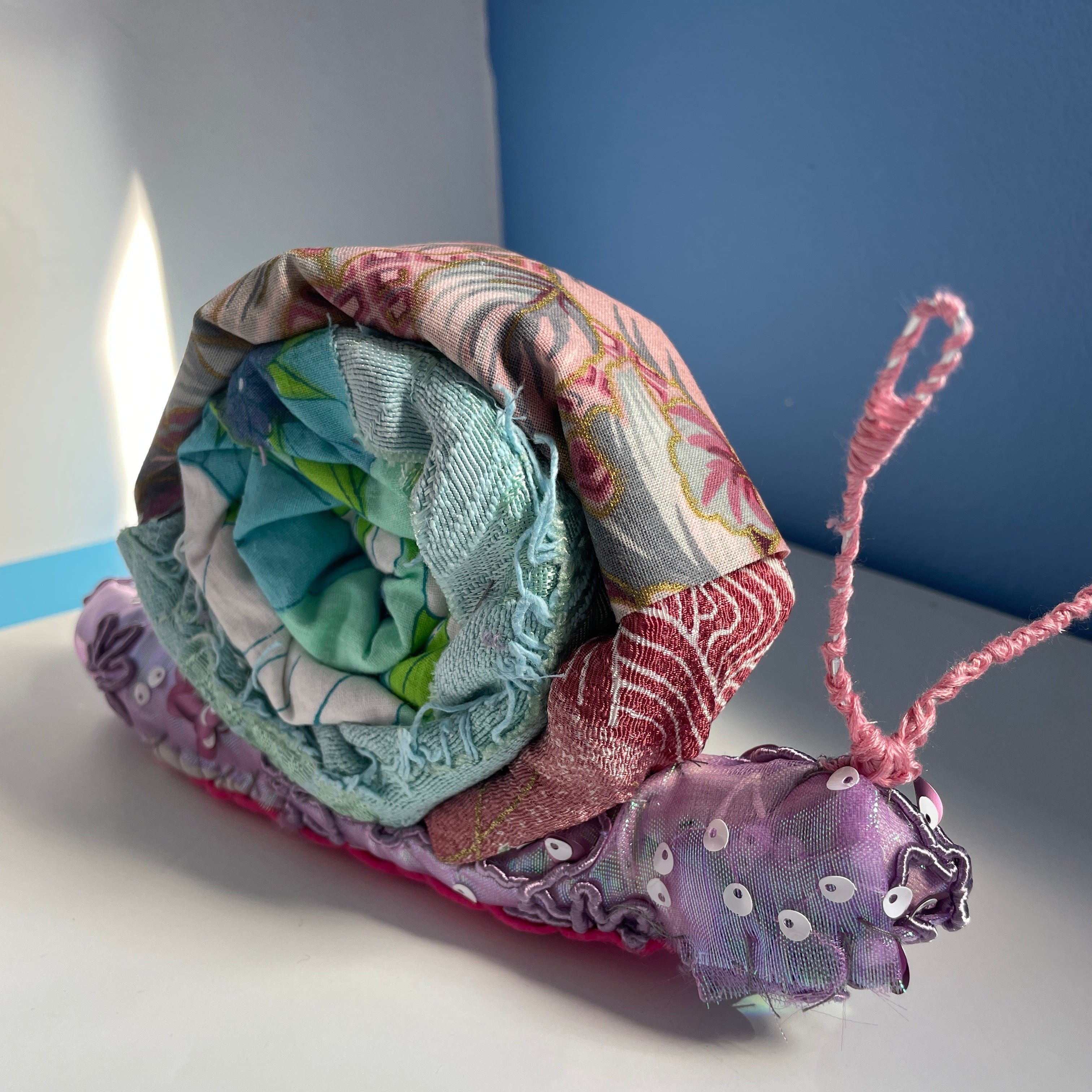 A handmade fabric snail decoration made from patterned and solid colored cloth layers, with pink yarn antennae and sequins on the body, resting on a white surface against a blue and white background.