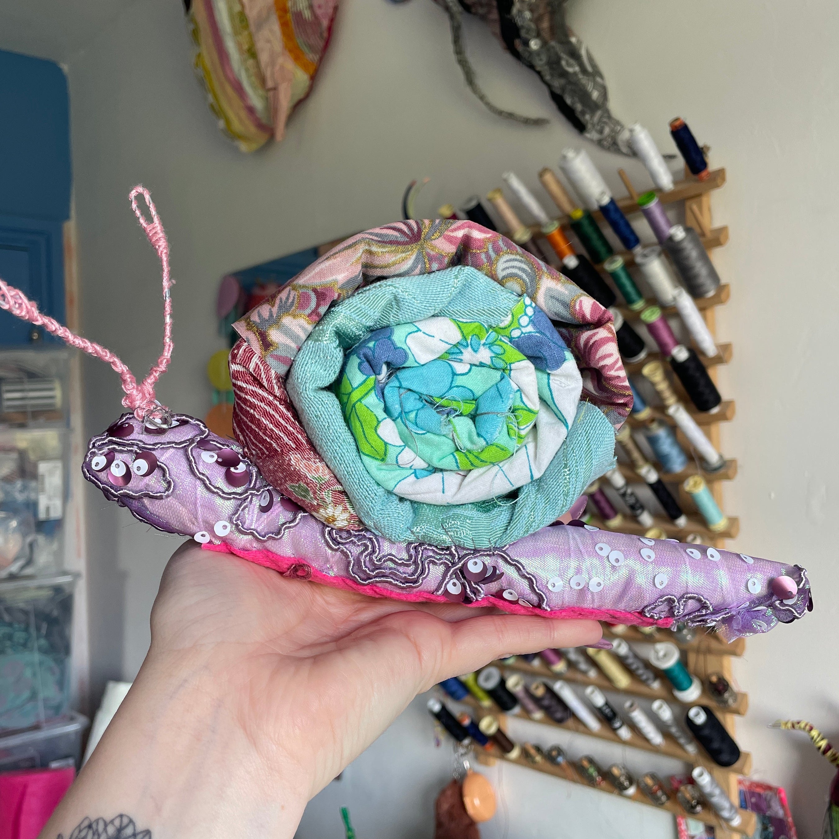 A hand holds a small handmade quilted fabric snail with a purple body, pink antennae, and a blue and green patterned shell in a craft room setting.