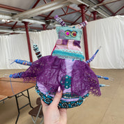 A handmade, highly textured insect-like fabric art piece being held by a hand in a warehouse setting. The piece features a purple lace body, teal and pink patterned fabric sections, and multiple blue and purple thread legs.