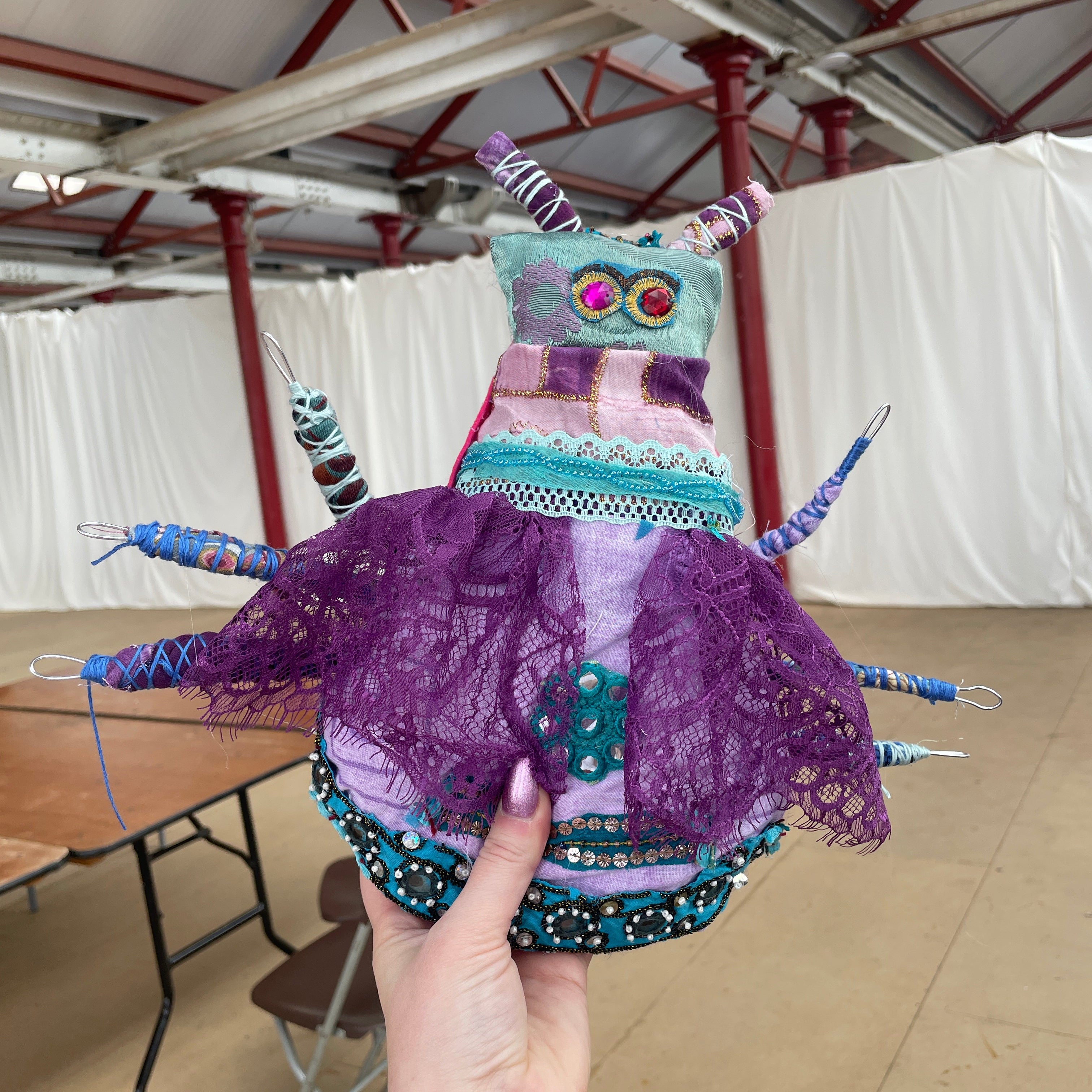A handmade, highly textured insect-like fabric art piece being held by a hand in a warehouse setting. The piece features a purple lace body, teal and pink patterned fabric sections, and multiple blue and purple thread legs.