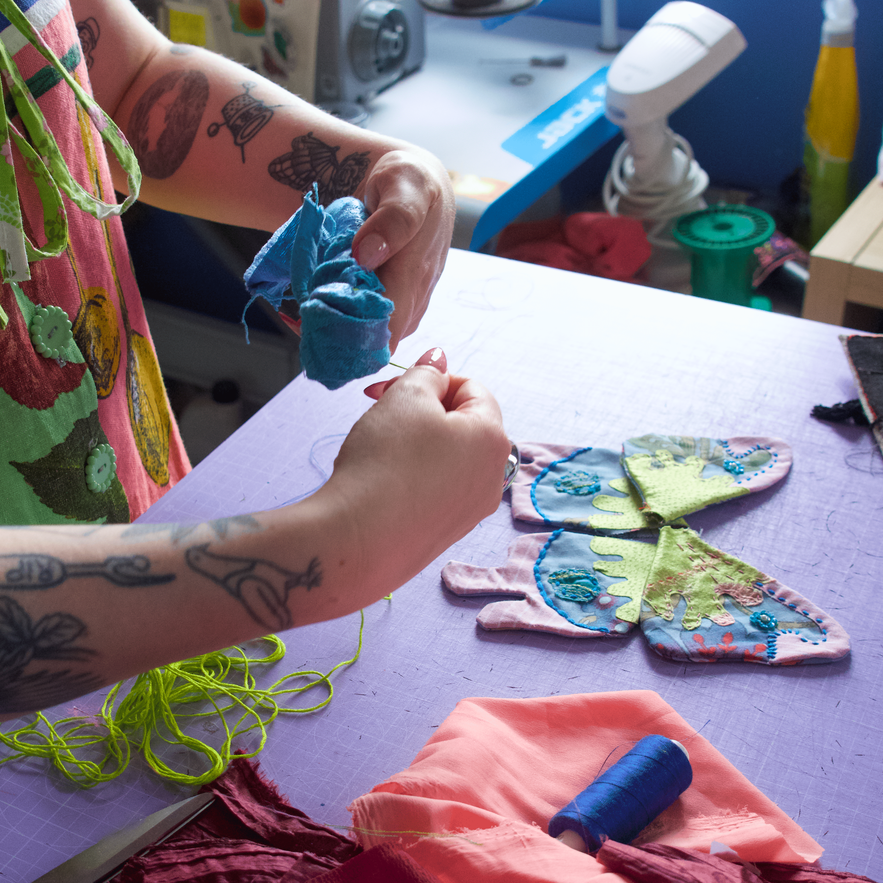A person with tattooed arms works at a lavender craft table, holding a small blue fabric piece. Several colorful fabric wings or appliqués with blue and green details are laid out on the table, alongside thread spools, pins, and other crafting supplies in a workshop setting.
