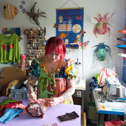 A woman with bright pink hair sits at a table in a cluttered, colorful sewing studio, working on a handmade quilt. The room is filled with sewing machines, shelves of fabric, and various colorful craft decorations.