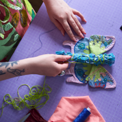 A close-up of a person's hands crafting a butterfly-shaped fabric item on a purple cutting mat, surrounded by sewing supplies including pins, thread, and a black fabric panel.