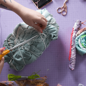 A top-down view of a person's hands crafting a quilted fabric item on a purple cutting mat, using orange-handled scissors to tie and trim a bundle of fabric. Other sewing tools, thread, and finished quilted pieces are visible around the workspace.