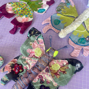 A handmade butterfly-shaped fabric ornament or plush toy made from multicolored patterned fabrics, including green, pink, purple, and black, with embroidered details and antennae, placed on a purple cutting mat alongside other butterfly crafts.