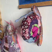 Two handmade fabric winged dolls or butterflies hanging on a white wall in a craft room setting. The right doll is made from a floral patterned fabric with purple, pink, and blue sections, and has a blue fabric wing.