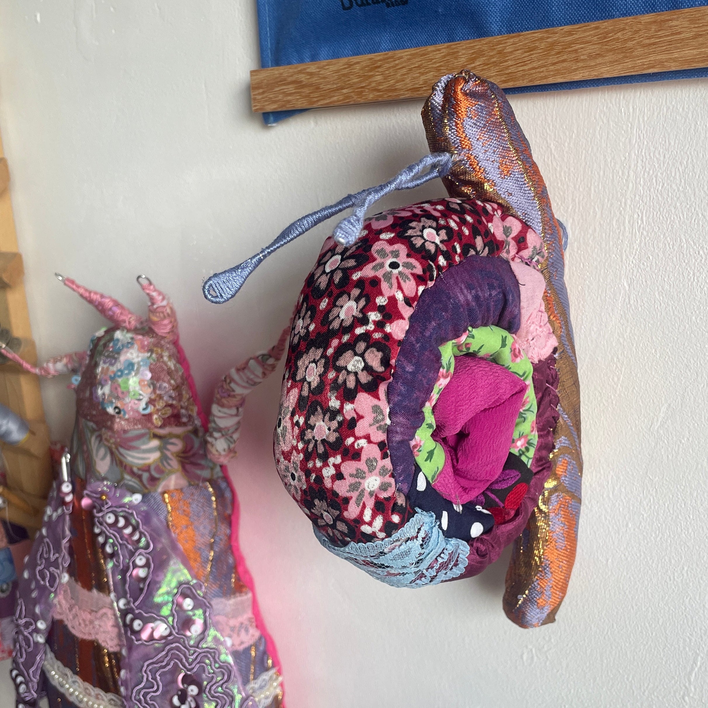 Two handmade fabric winged dolls or butterflies hanging on a white wall in a craft room setting. The right doll is made from a floral patterned fabric with purple, pink, and blue sections, and has a blue fabric wing.
