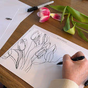 Person drawing tulips on paper with a pen, next to actual tulips on a wooden table.