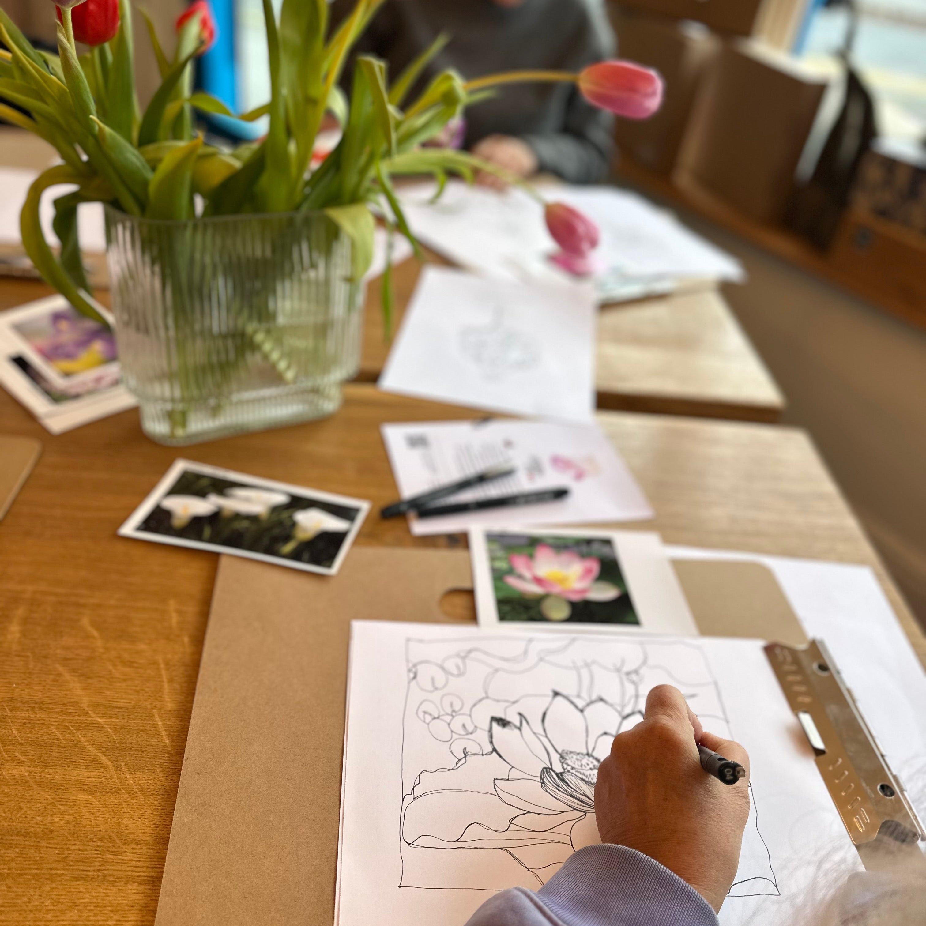 Person drawing a flower on paper with flowers and stationery items on a table.