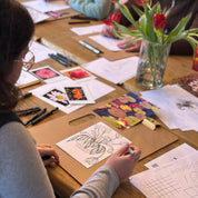 People working on art projects at a table with flowers and stationery.