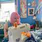 Person with pink hair using a sewing machine in a room with blue walls and various decorations.