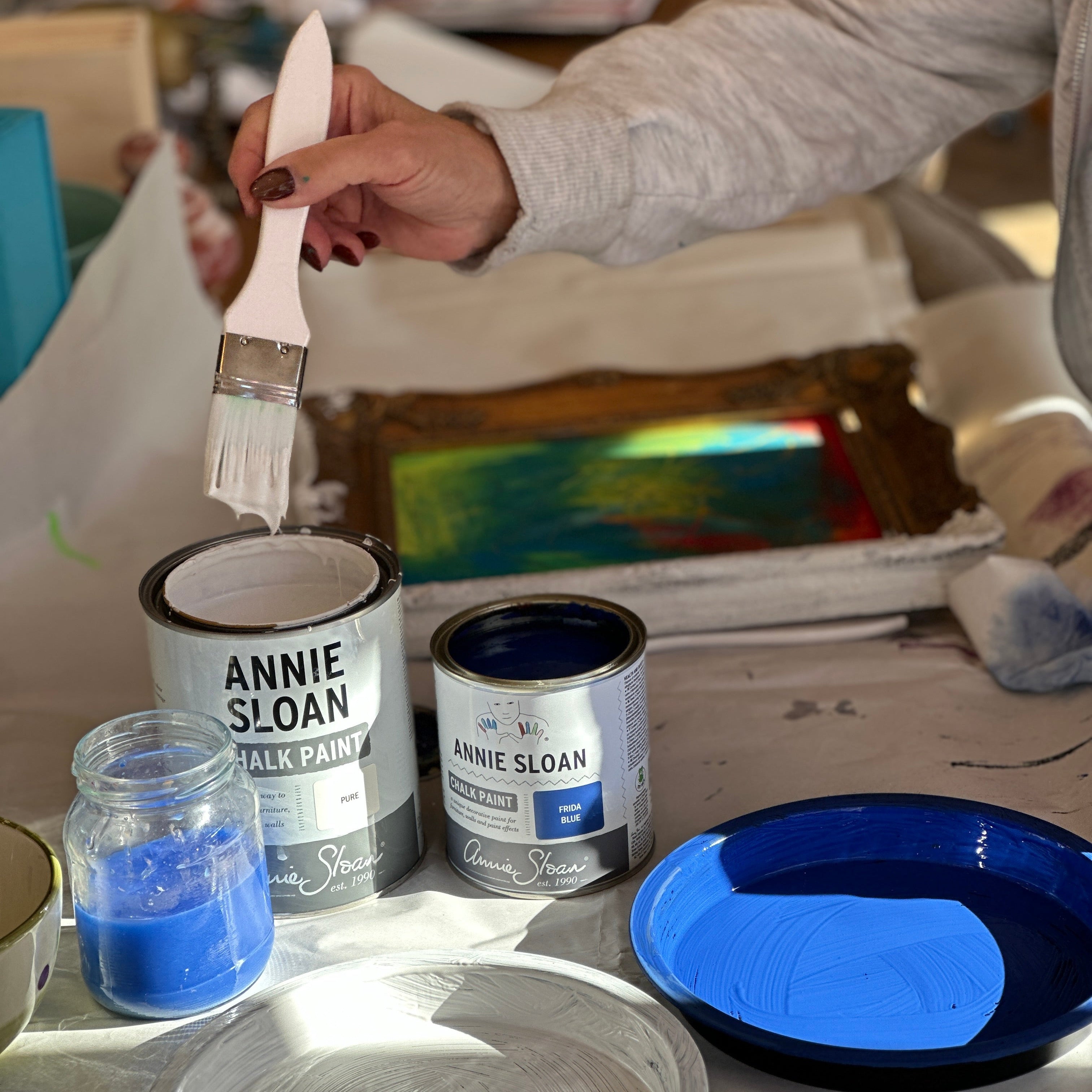 Person painting a surface with Annie Sloan chalk paint, surrounded by paint cans and a blue plate.
