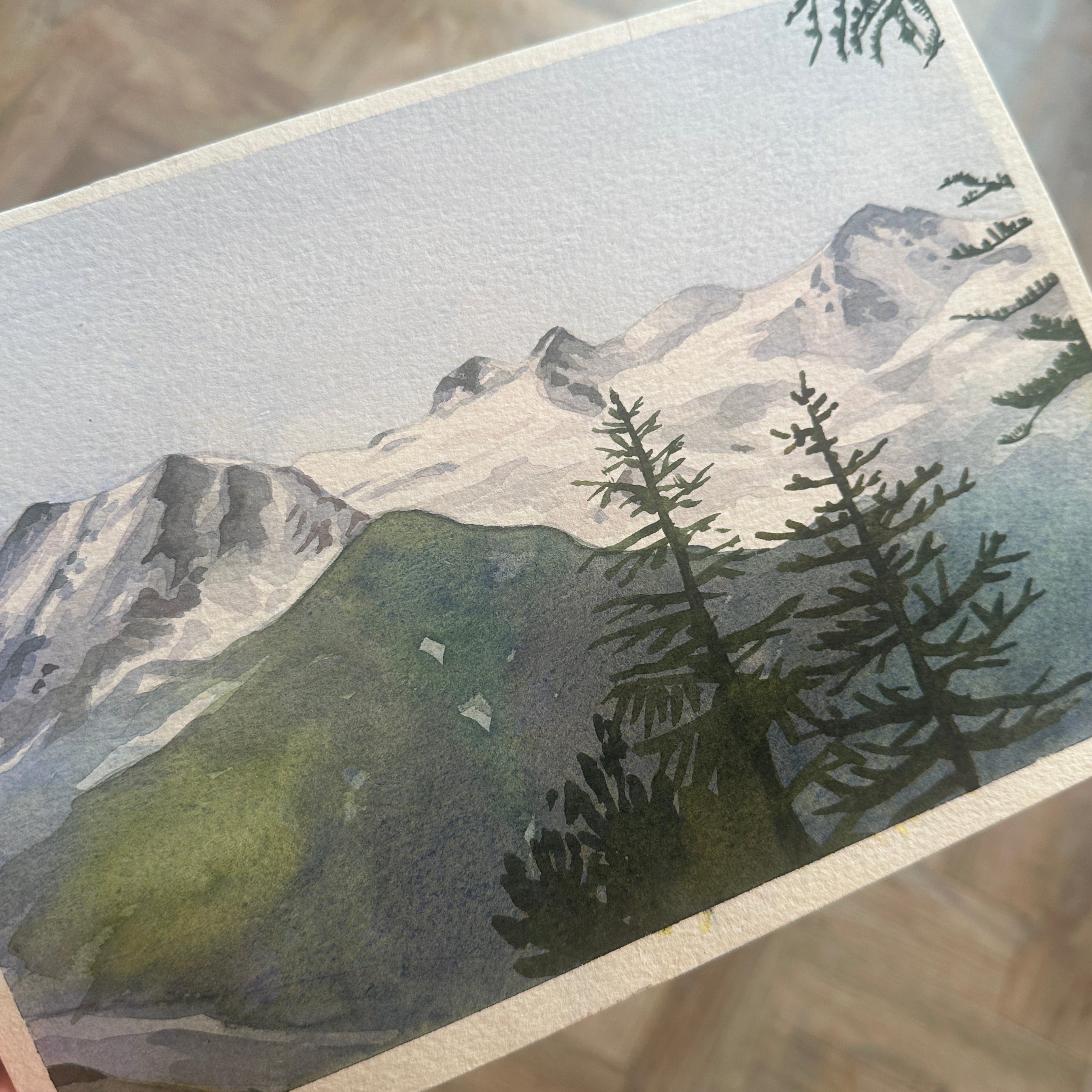Hand holding a card with a mountain and tree illustration on a wooden surface