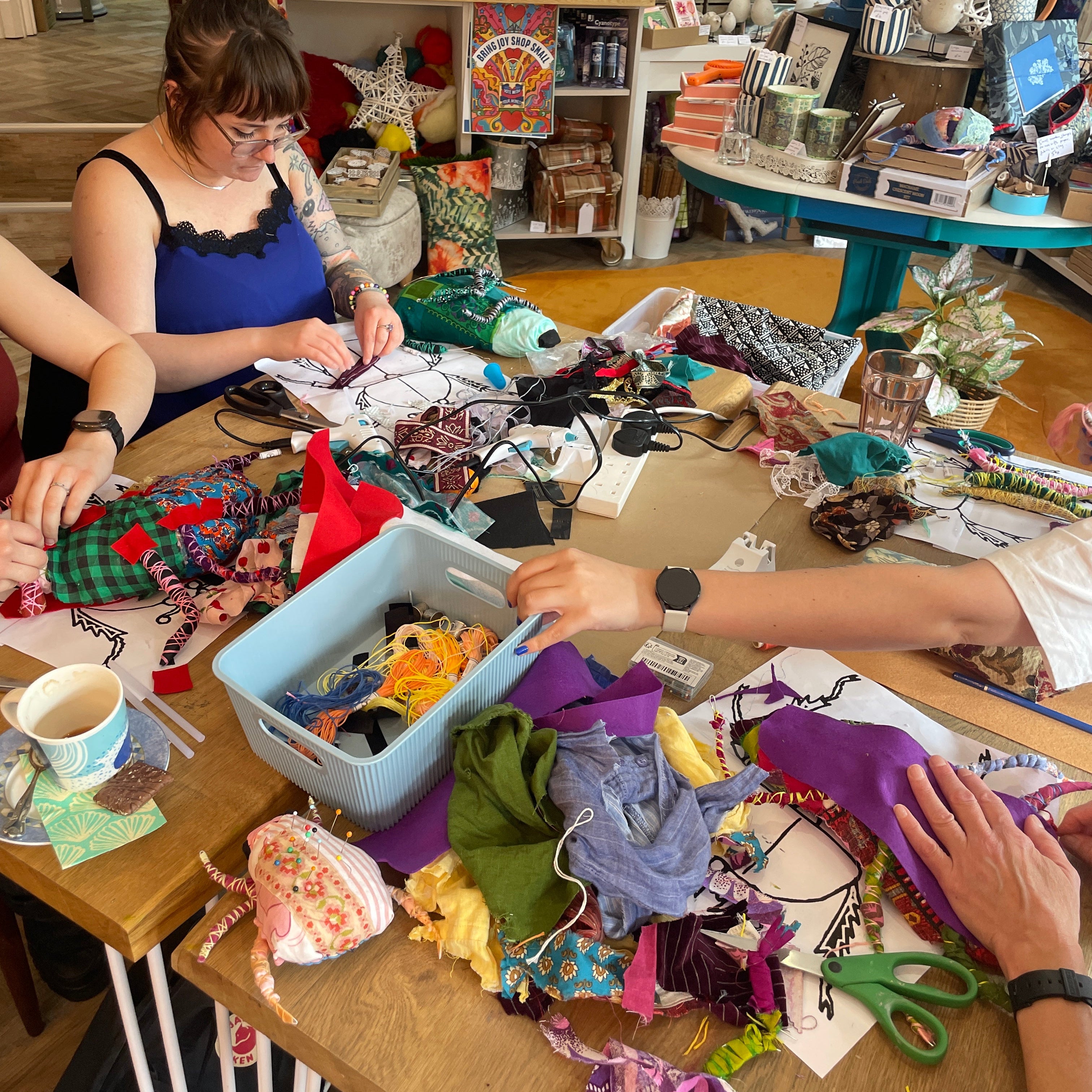 A busy craft or sewing workshop scene with multiple people visible from the shoulders down, working at a wooden table covered with fabric, scissors, notions, and a light blue sewing machine basket. The environment is cluttered but clearly a real indoor workspace.