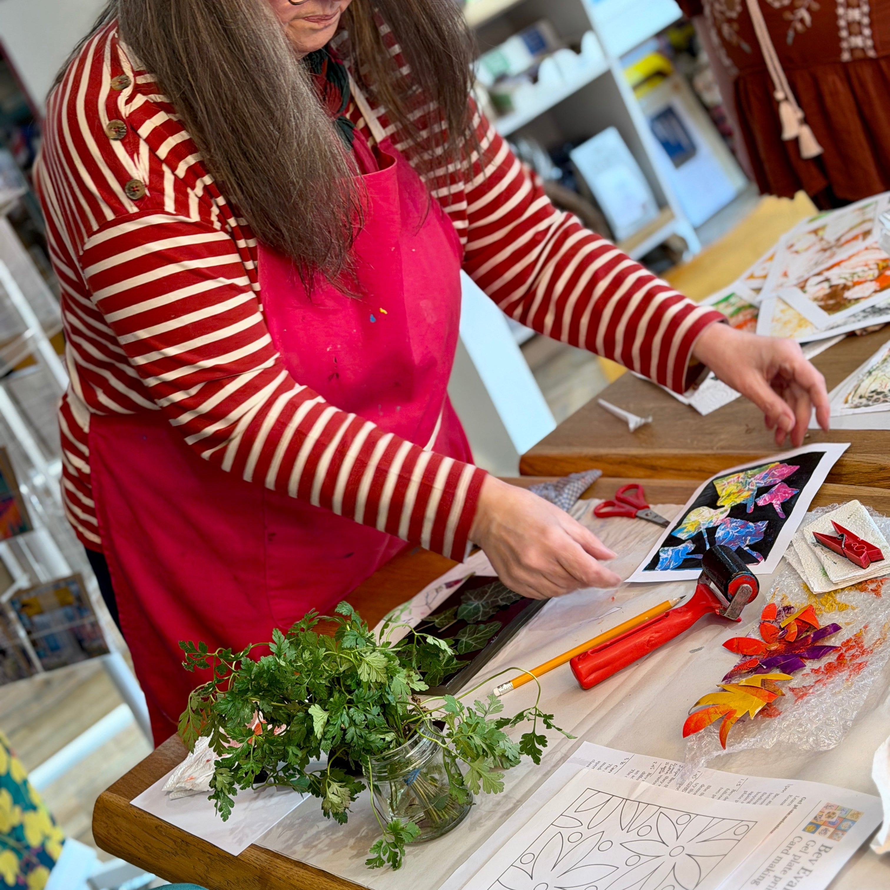 Woman in a red apron working at a table with art supplies in a classroom setting