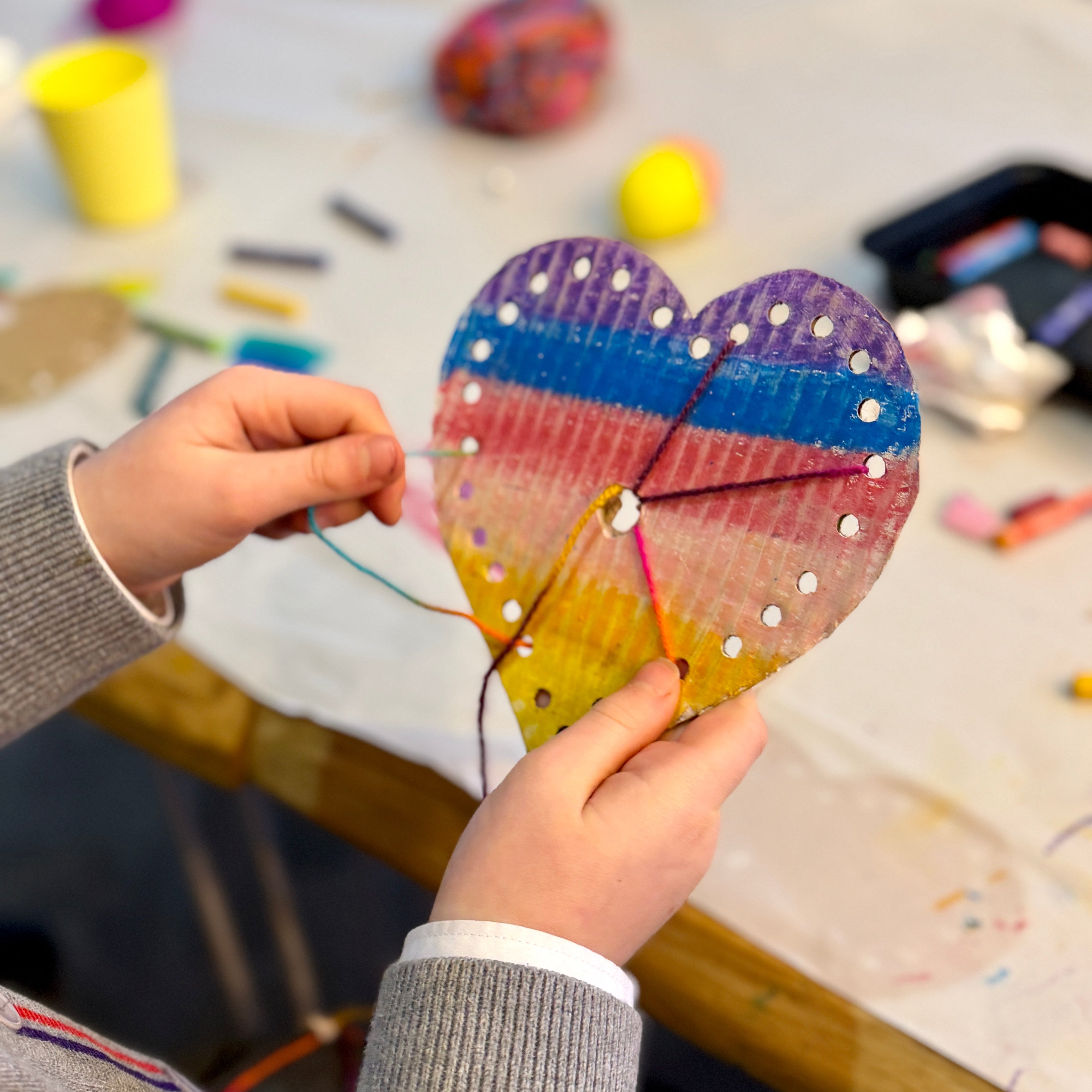 Child's hands holding a colorful heart-shaped craft with a blurred art studio background.