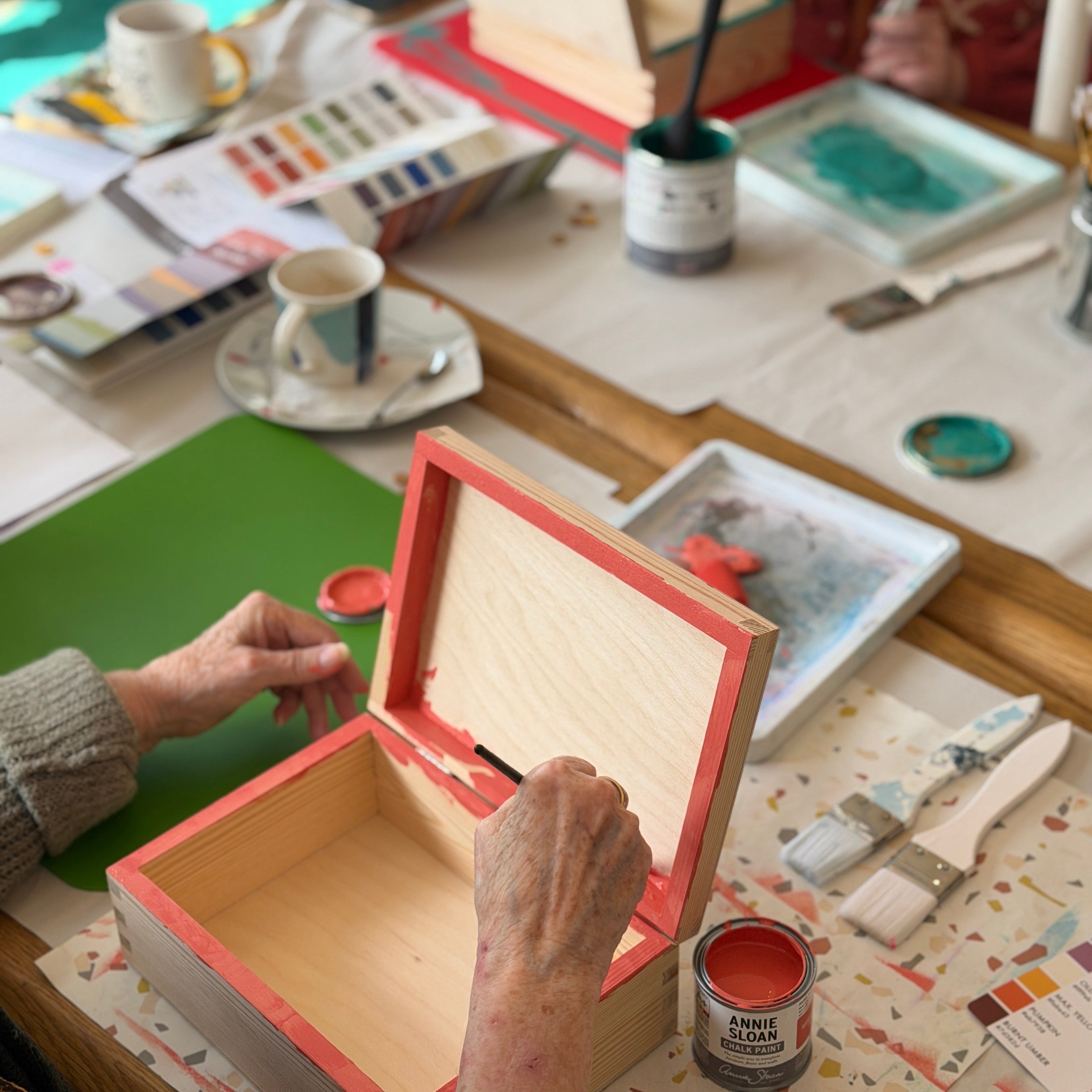 Person opening a wooden box on a table with art supplies and a cup of coffee.