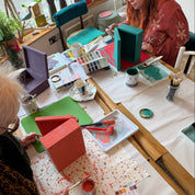 Two people working on small wooden houses at a table with art supplies.