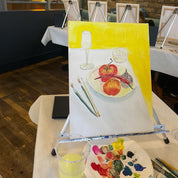 A still life painting on canvas showing apples on a plate, a wine glass, and paintbrushes, displayed on an easel in a studio or workshop setting with other easels and a paint palette in the foreground.