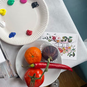 An overhead view of a tabletop setup with a white paper plate holding painted paint dollops and another plate holding a tangerine, a red chili pepper, and a dark purple eggplant. A floral art print and part of a person's legs in metallic shoes are also visible on a white tablecloth.