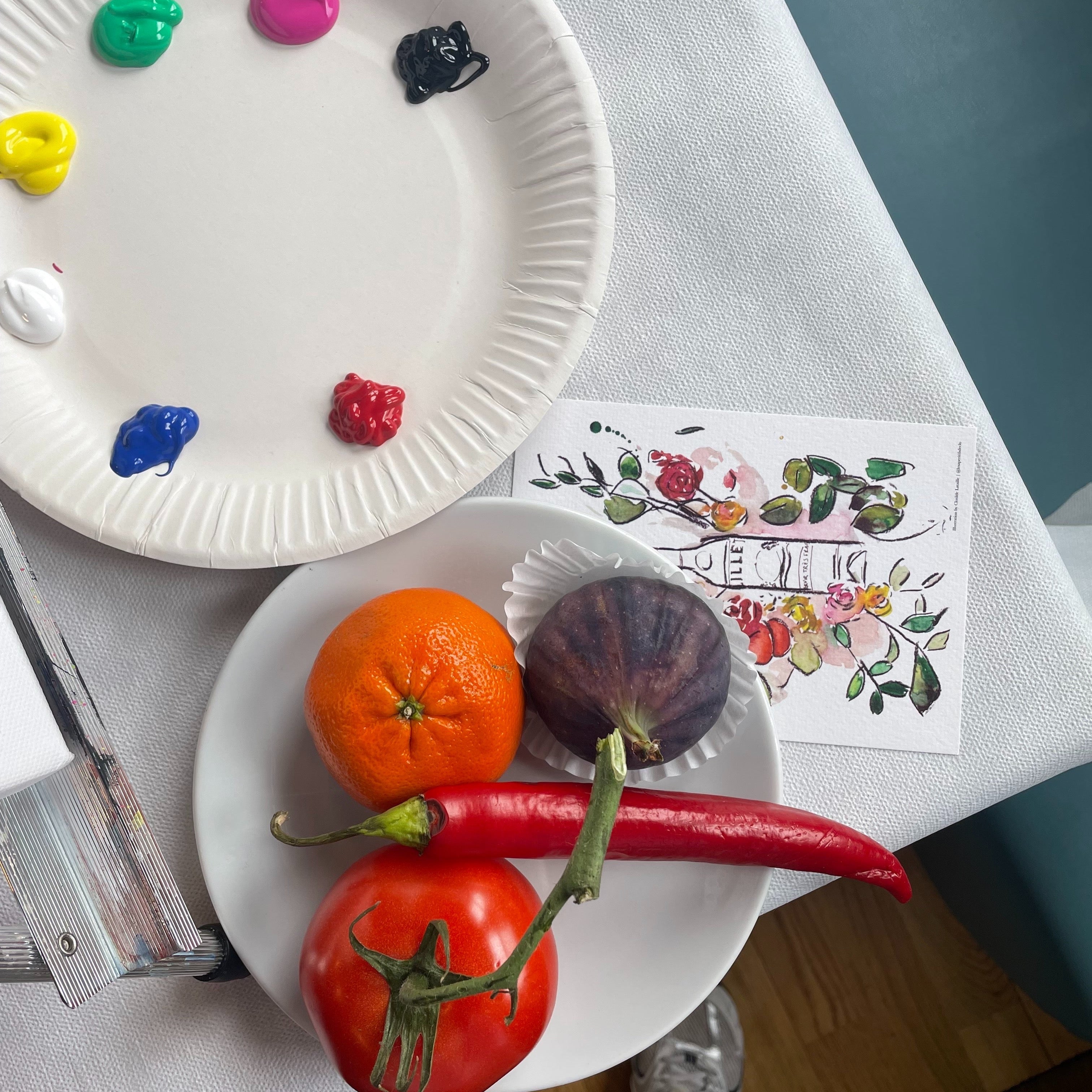 An overhead view of a tabletop setup with a white paper plate holding painted paint dollops and another plate holding a tangerine, a red chili pepper, and a dark purple eggplant. A floral art print and part of a person's legs in metallic shoes are also visible on a white tablecloth.