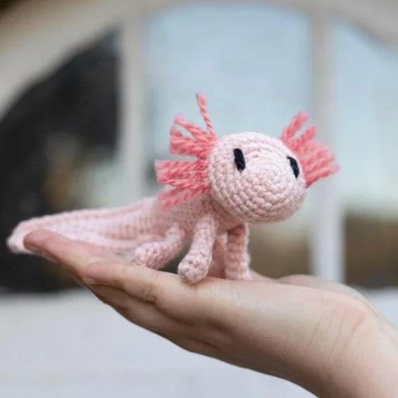 Pink crochet axolotl toy held in a hand with a blurred background