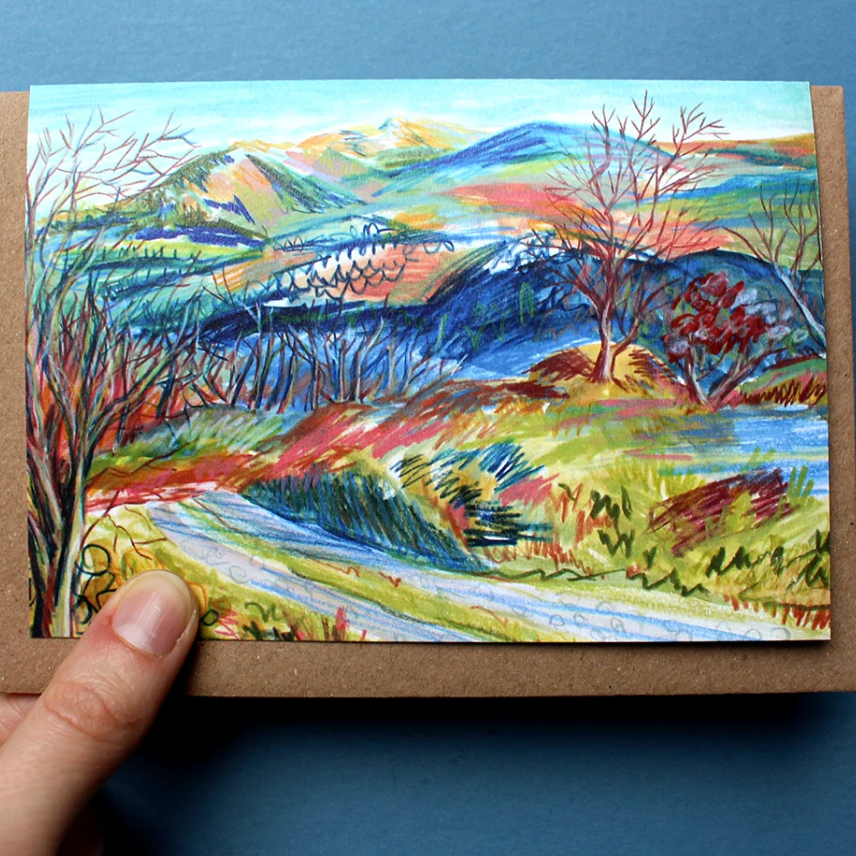 A hand holds a small sketch art card featuring a colorful landscape with blue mountains, green fields, and red trees, shown on a webpage next to product details for a North Wales sketch card set.