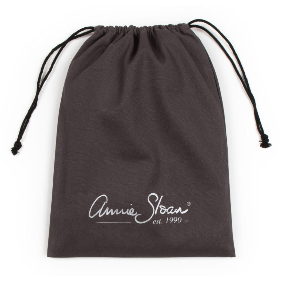 A screenshot of an e-commerce product page for the Annie Sloan Apron, showing a dark brown apron with a white script logo and a matching dark brown drawstring bag on the right. The left side contains product details, pricing, and a pink 'Kiarna available at checkout.