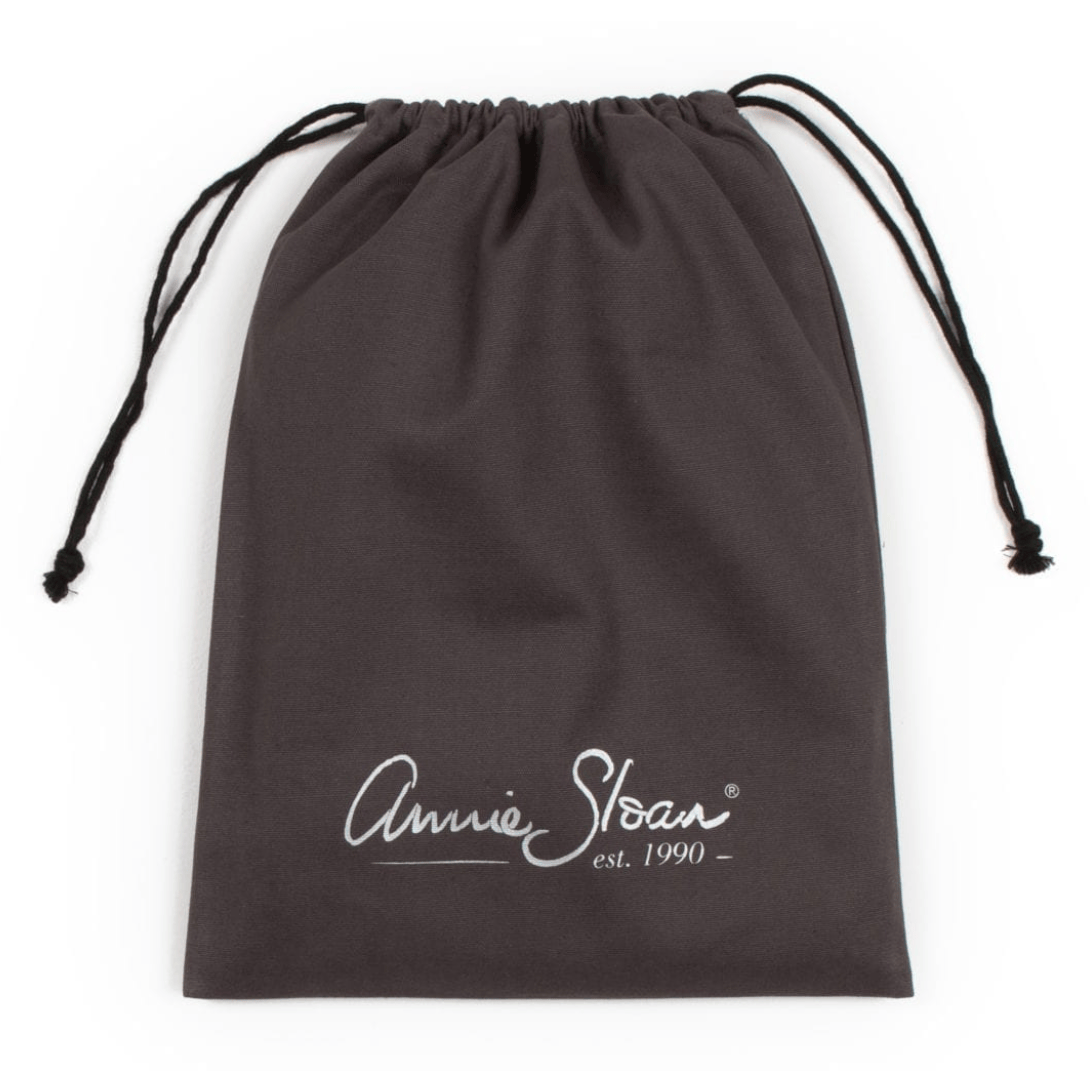 A screenshot of an e-commerce product page for the Annie Sloan Apron, showing a dark brown apron with a white script logo and a matching dark brown drawstring bag on the right. The left side contains product details, pricing, and a pink 'Kiarna available at checkout.